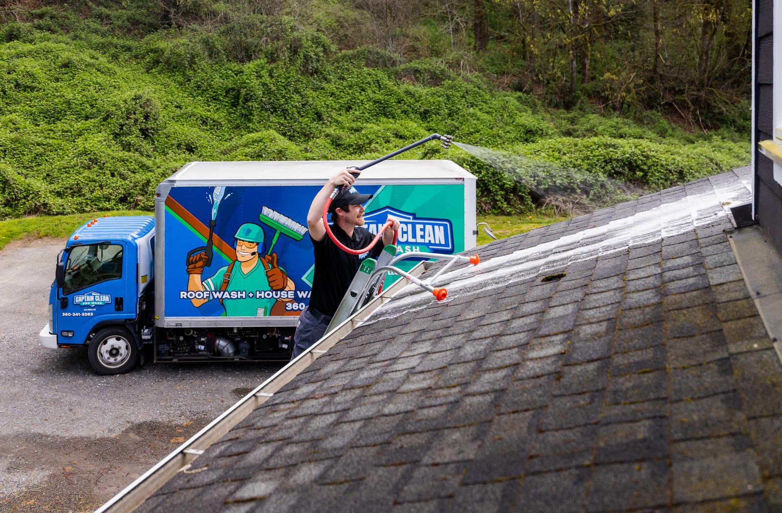 Captain Clean Pro Wash technician applying roof treatment to moss-stained shingles beside the branded service truck